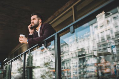Resmi takım elbiseli ciddi etnik bir adamın yeni bir içecek içmesi, iş arkadaşıyla konuşması, balkonda çitlere yaslanarak başka yere bakması.