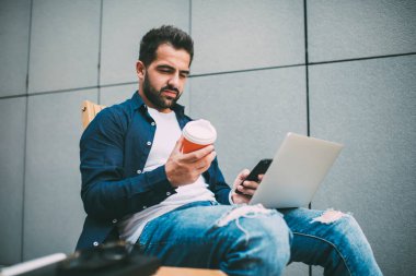Kafkasyalı, tek kullanımlık kahve fincanı hakkında bilgi okuyan ciddi bir beyaz erkek. Cihazlar aracılığıyla geri bildirim okuyor. Kafedeki dizüstü bilgisayar dersinde ders alırken düşünceli bir erkek öğrenci ara veriyor.