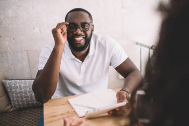 Göz koruyucu gözlüklü neşeli Afro-Amerikan adamın portresi özel ders sırasında kameraya gülüyor üniversite projesinde işbirliği için, esmer tenli erkek ve kız kafede.
