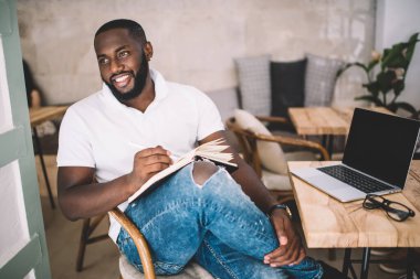 Eğitim kitabıyla çalışan koyu tenli mutlu bir öğrenci boş dizüstü bilgisayarla masanın yanında oturuyor ve öğrenme zamanının tadını çıkarıyor. Üniversite web sitesi için fotokopi alanı olan modern bir bilgisayar.