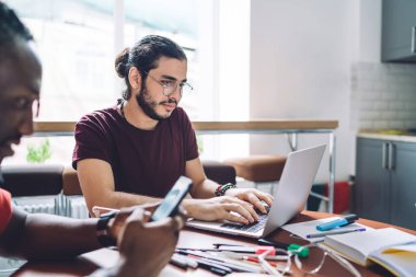 Gözlüklü ve günlük giysiler içinde çalışkan genç bir çalışan ekranda yoğunlaşırken ve netbook ile etkileşime girerken aynı zamanda ekin dalgasındaki Afro-Amerikan iş ortağının yanında otururken akıllı telefon kullanıyor.