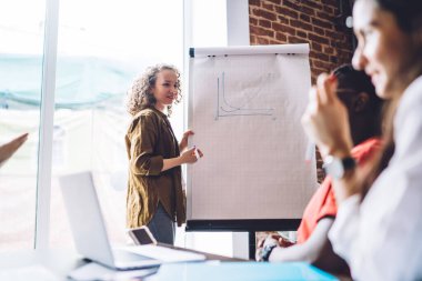 Gündelik giyinen genç ve zeki bir bayan modern ofiste toplantı yaparken çalışma arkadaşlarına grafikleri gösteriyor. 