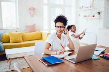 Küçük kız tabletle oynarken ve gülümserken, dizüstü bilgisayarının başında oturup telefonla konuşan mutlu Afrikalı Amerikalı anne.