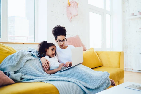 少数族裔母亲戴着眼镜 与可爱的孩子坐在蓝色毛毯下的黄色沙发上 同时在笔记本电脑上观看社交媒体的视频 — 图库照片