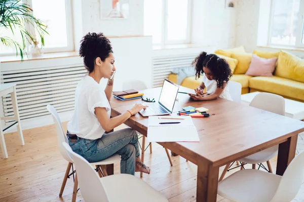 Genç Afrikalı Amerikalı kadın cep telefonuyla konuşurken boş ekranla çalışırken çalışma odasında küçük bir kızla modern evde sörf tabletinde otururken yan görünüm. 
