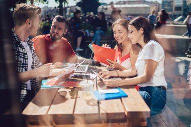Bir grup genç arkadaş belgelere bakıyor ve açık kafede oturmuş güneşli bir günde ödev yaparken verileri tartışıyorlar.