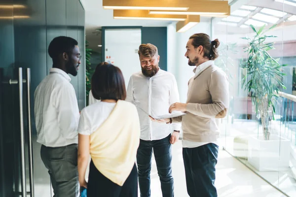 Group of successful multiracial colleagues standing in office and discussing business project during cooperation indoors, confident diverse team 20 year old enjoying live communication at workspace