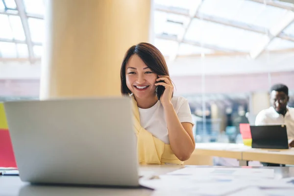 Asian woman smiling during cellphone conversation in modern office interior, happy Japanese female professional discussing ideas for project using mobile phone for calling and laptop for info browsing