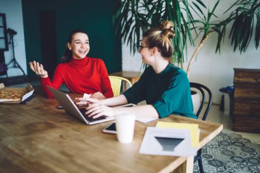 Keyifli genç bir kadın dizüstü bilgisayar kullanıyor ve masada oturup ev ödevlerini tartışırken mutlu arkadaşına bakıyor.