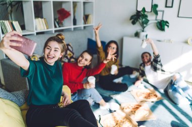 Keyifli arkadaşlar günlük giysiler içinde lezzetli pizzalar yiyor ve odada dinlenirken kahveden zevk alıyorlar ve bulanık arka planda selfie çekiyorlar.