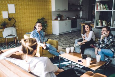 Bir grup neşeli genç öğrenci küçük renkli bir modern tasarım dairesinde oturuyor. Kahve, dizüstü bilgisayar, akıllı telefonlar ve birbirleriyle konuşuyorlar.