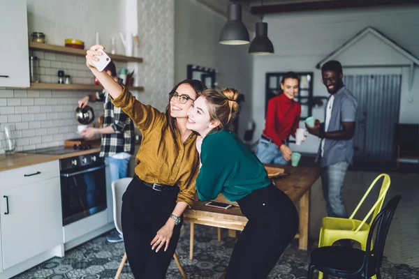 Delighted women in casual clothes hugging and taking photo with smartphone while multiethnic people preparing for meal in kitchen in urban apartment