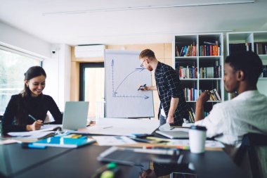 Modern ofisteki çok ırklı meslektaşlar için veri sunumu yaparken günlük kıyafet çizimi yapan genç bir erkek parmak izi grafiğinde büyüyen bir tablo çiziyor.