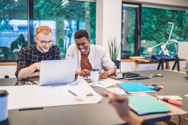 Dizüstü bilgisayarla eğitim projesi hazırlarken gülen ve kütüphanedeki masada kahve içen genç grup arkadaşları.