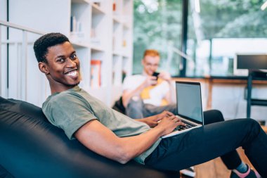 Afro-Amerikan bir adamın neşeli bir şekilde gülümsemesi ve kameraya bakması. Fasulye torbasının üzerinde otururken ve yaratıcı çalışma ortamında dizüstü bilgisayarda klavye yazarken.