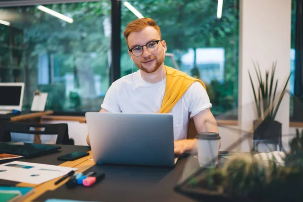 穿着休闲装的白色T恤的快乐聪明的红头发男性坐在笔记本电脑前浏览 桌上放着一大杯背景咖啡 — 图库照片