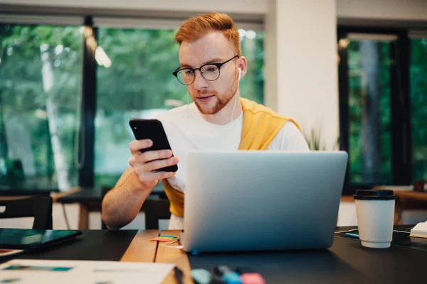 年轻而专注的红头发男性 身穿休闲装 穿着白色T恤 在手机上浏览着 坐在桌旁 端着咖啡和笔记本电脑 — 图库照片
