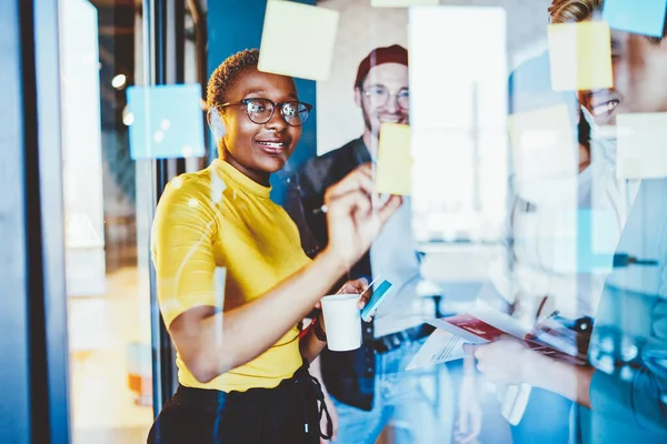 Group of happy hipster guys making notes on colorful memo sticks at glass wall while creating interesting ideas, cheerful multiracial male and female smiling while working with startup project