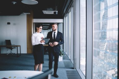 Ofis kıyafetleri içinde elinde tablet olan neşeli bir kadınla konuşurken ofis koridorunda pencerenin yanında duruyor.