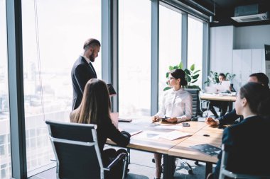 Elinde kağıt olan odaklı bir adam ofis binasında toplantı sırasında masada dizüstü bilgisayarla oturan çalışanların önünde duruyor.