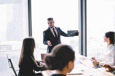 Modern ofis binasının penceresinde dikilirken şık saç kesimi olan sakallı iş adamı yönetim kuruluna tabloyu sunuyor.