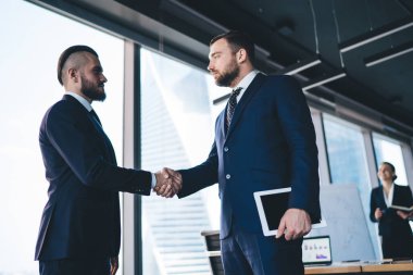 Sakallı, resmi takım elbiseli, düşünceli bir işadamından. Büyük pencerelerin yanında duran ciddi bir ortağın elini sıkıyor.