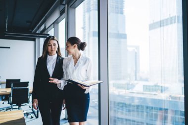 Büyük defterli kadın asistan, şehir merkezindeki modern ofis binasının penceresinin yanında dikilirken etnik iş kadınıyla konuşuyor.