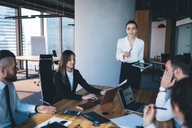 Kadın elinde tabletle ofiste duruyor ve iş arkadaşları laptoplarla masada oturup grafiklere bakarken ve soru sorarken rapor yazıyor.
