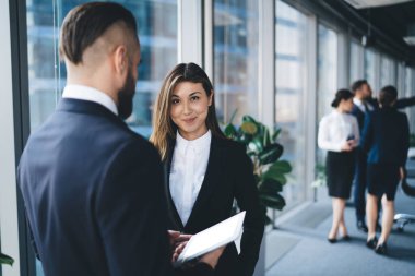 Takım elbiseli, güler yüzlü kadın resmi giyinmiş kendine güvenen bir adamın yanında duruyor. Belediye binasının penceresinin yanında tablet taşıyor.