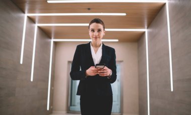 Katı takım elbiseli, şık ve neşeli kadın çalışan. Parlak koridorda duruyor ve kameraya bakarken sosyal medyada mesajlaşıyor.