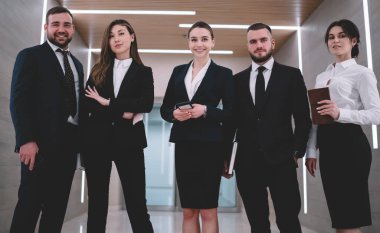 Katı takım elbiseli saygıdeğer ve mutlu profesyonellerden oluşan bir grup koridorda yaratıcı lambalarla durup kameraya bakarken gülümsüyor.