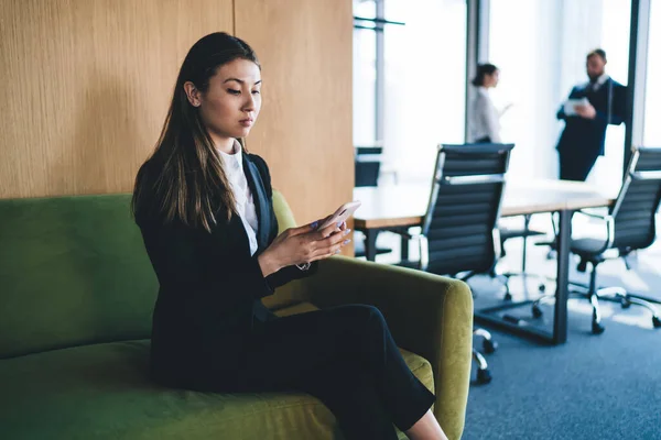 年轻的女经理穿着雅致的西装 在办公室的模糊背景下 坐在舒适的沙发上 一边浏览智能手机 — 图库照片