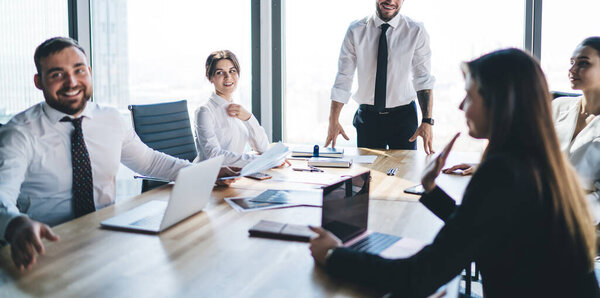 Веселые сотрудники в формальной одежде сидят за столом, обсуждают рабочие вопросы, улыбаются и слушают босса урожая, стоящего у окна в офисе