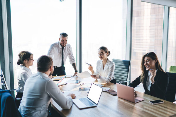 Руководитель группы, стоящий и говорящий о процессе работы со смехом, в то время как коллеги сидят за столом и анализируют документы в легком офисе 