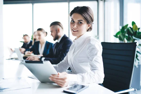 Smart formal woman working with tablet at table with group of colleagues and looking at camera with smile in light office