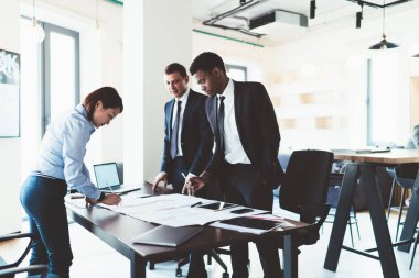 Çok ırklı genç iş arkadaşları ellerinde cihazlar ve belgelerle masanın yanında duruyor ve şirket bürosunun toplantı odasında proje üzerinde çalışıyorlar.