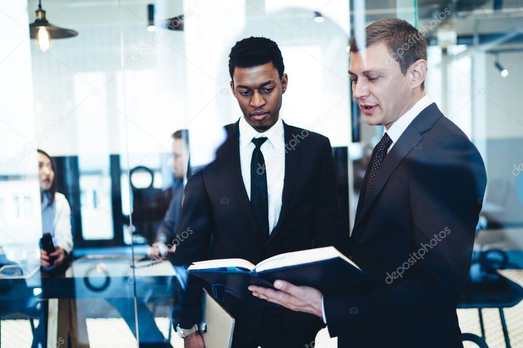 Through glass view of director in business style suit standing and announcing plan for African American employee while holding and looking at appointment book