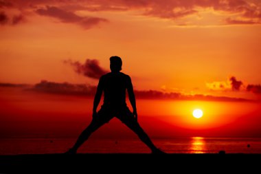 Silhouette of male athlete working out cross training on colorful sunrise background, beautiful silhouette of sportsman doing stretching exercise standing on the beach clipart