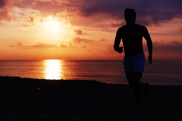 Silhueta de atleta de construção muscular correndo rápido um log na praia, corredor em ação correndo contra o nascer do sol colorido sobre o mar, corredor masculino com corpo muscular em ação — Fotografia de Stock