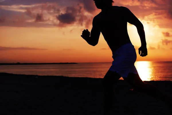 Silueta de la construcción muscular atleta corriendo rápido un registro de la playa, corredor en acción trotando contra el colorido amanecer sobre el mar — Foto de Stock