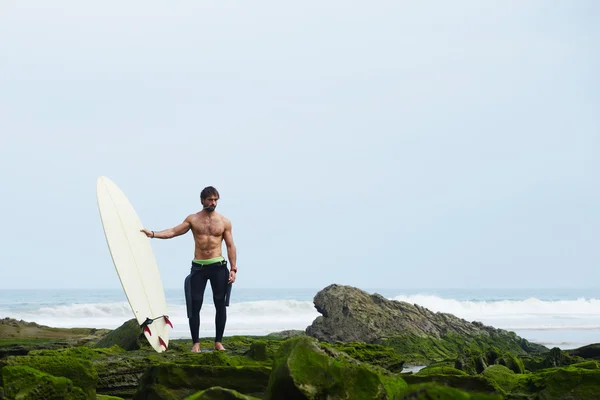 Professional surfer in black wetsuit standing on moss rocks — Stock Photo, Image