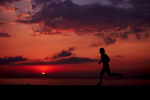 Perfecta y hermosa silueta de hombre deportivo en forma corriendo en la playa, entrenamiento de fitness matutino al aire libre, joven corredor corriendo rápido al amanecer colorido —  Fotos de Stock