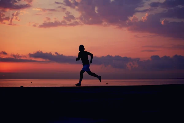 Perfecta y hermosa silueta de hombre deportivo en forma corriendo en la playa, entrenamiento de fitness matutino al aire libre, joven corredor corriendo rápido al amanecer colorido —  Fotos de Stock