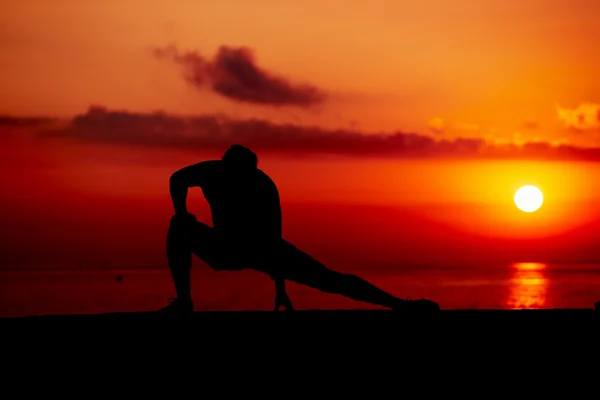 Silhuett av ung man på terränglöpning under färgglada sunrise på stranden, atletisk löpare med muskulös kropp gör stretching ben träning utomhus, fitness och hälsosam livsstilskoncept — Stockfoto