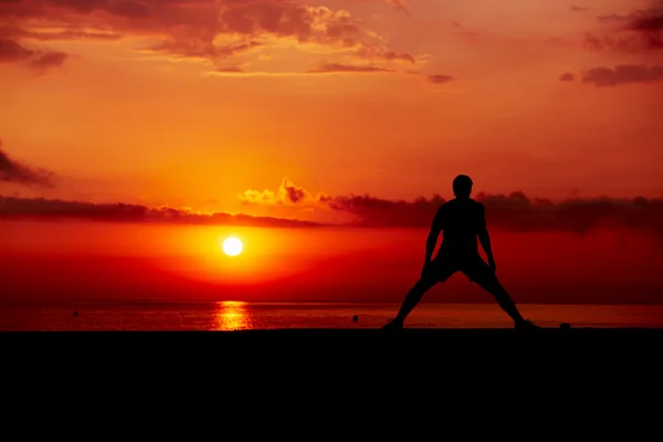 Silhouette d'athlète masculin s'entraînant en cross training sur fond de lever de soleil coloré, belle silhouette de sportif faisant des exercices d'étirement debout sur la plage — Photo