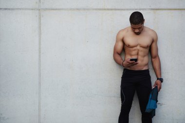 Handsome male sportsman with sexy muscular body using mobile phone with copy space area on button wall as side, runner chatting on smartphone outdoors