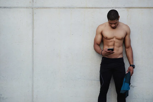 Handsome male sportsman with sexy muscular body using mobile phone with copy space area on button wall as side, runner chatting on smartphone outdoors — Stok fotoğraf