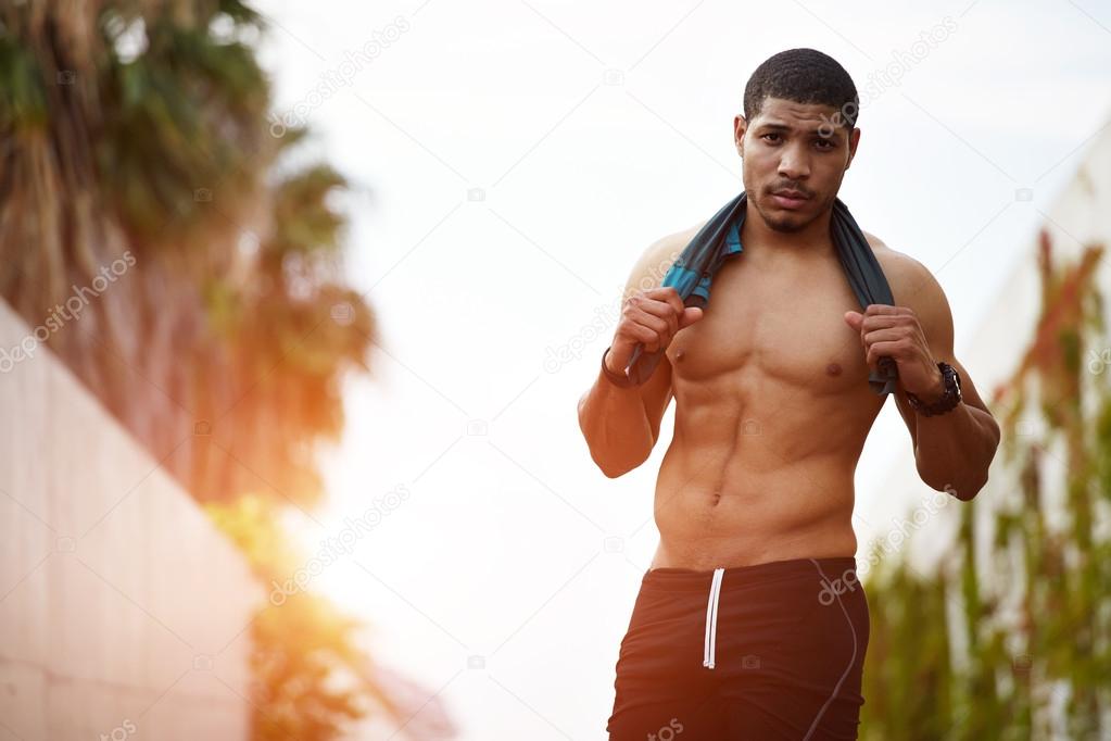 Portrait of dark skinned man with sexy abs resting after intense run outdoors while holding a shirt around his neck