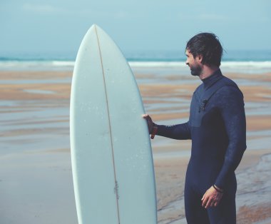Attractive young surfer holding surfboard while standing on the beach looking at ocean to find the perfect spot to go surfing waves,professional surfer with surf board looking at ocean, filtered image clipart