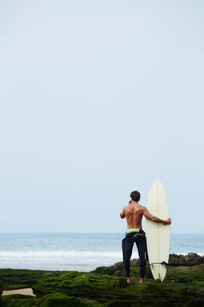 Oung surfista tenendo tavola da surf mentre in piedi sulla spiaggia e guardando l'oceano per trovare il luogo perfetto per andare onde surf, surfista professionista con tavola da surf guardando l'oceano, immagine filtrata — Foto Stock